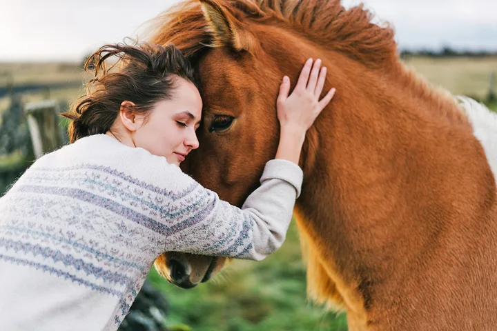 Young woman leaning her head tenderly against a chestnut horse's face in an outdoor pasture setting.