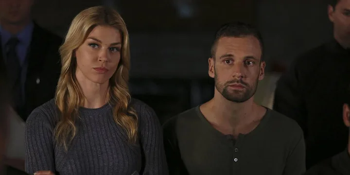 A blonde woman and a dark-haired man stand together looking serious in a dark indoor setting.