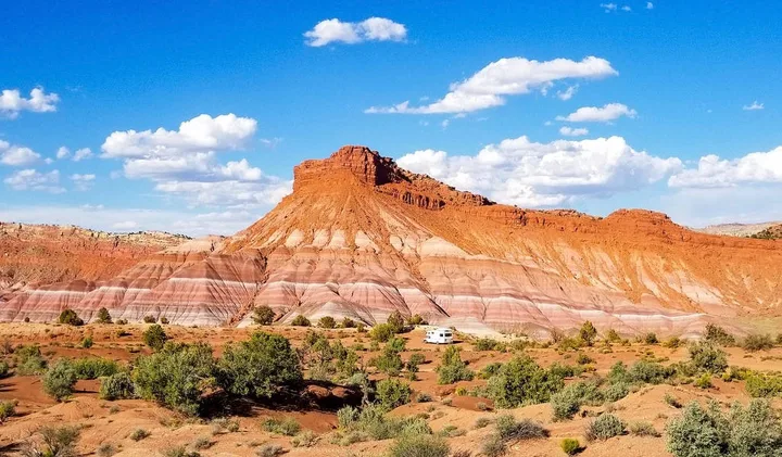 A white RV parked at the base of a massive red sandstone butte in a desert landscape.