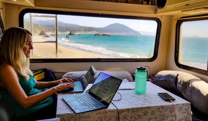 Woman working on a laptop inside an RV with a stunning ocean beach view through the window.
