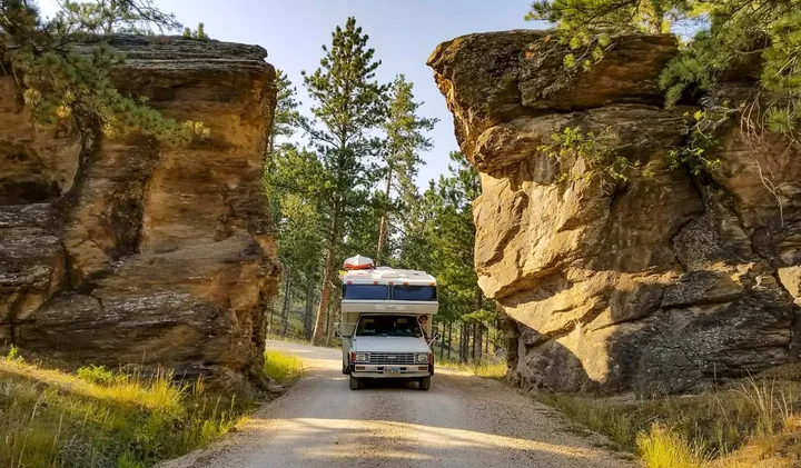 An RV driving through a narrow gap between towering sandstone rock formations on a dirt road.
