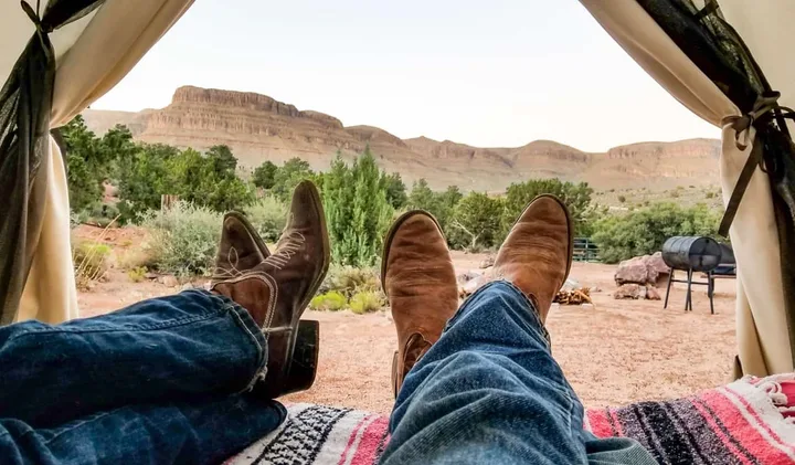 Two people relaxing in a tent doorway with cowboy boots, overlooking a desert canyon campsite.