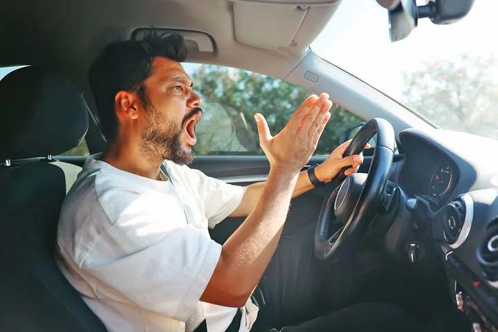 A frustrated man shouting and gesturing angrily while driving a car.