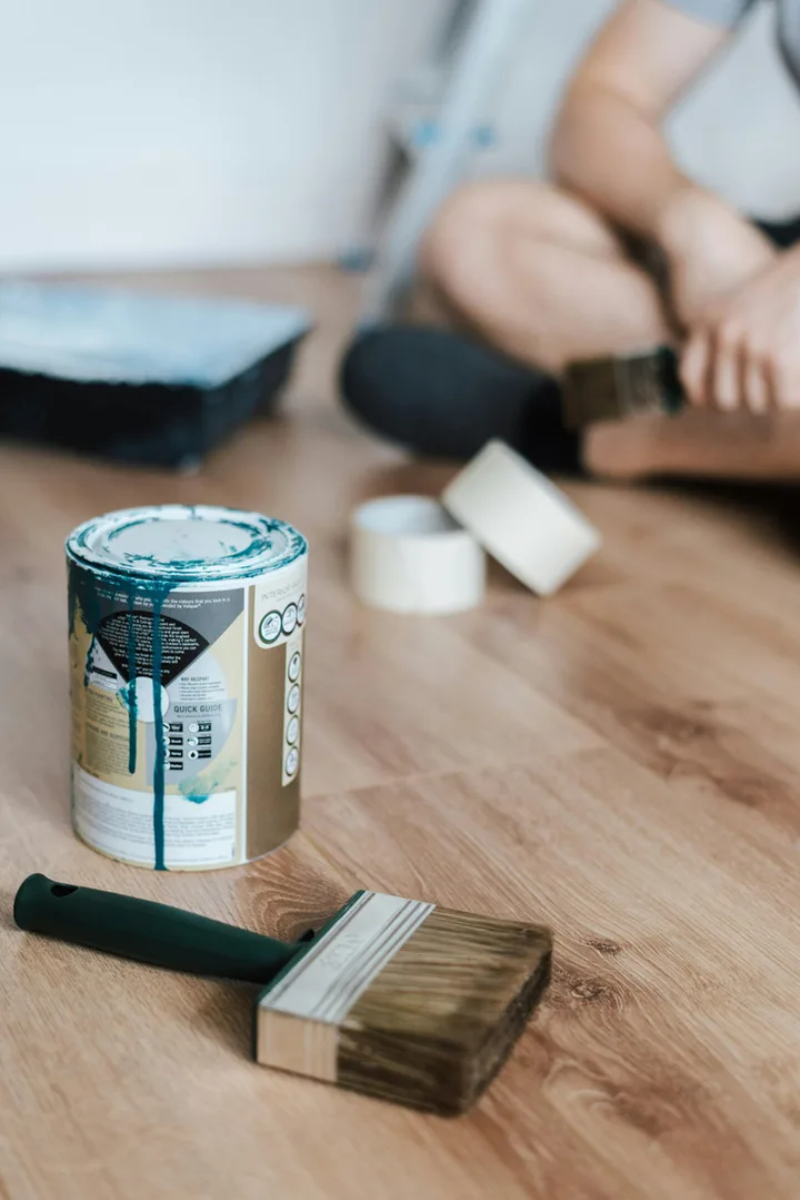 Paint can with dripping teal paint, a brush, and tape on a wooden floor during home renovation.