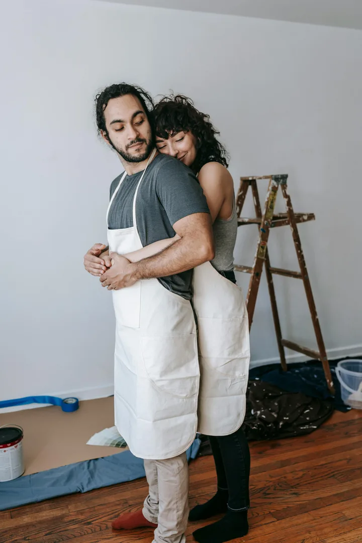 Couple in white aprons hugging affectionately inside a room being painted, ladder in background.
