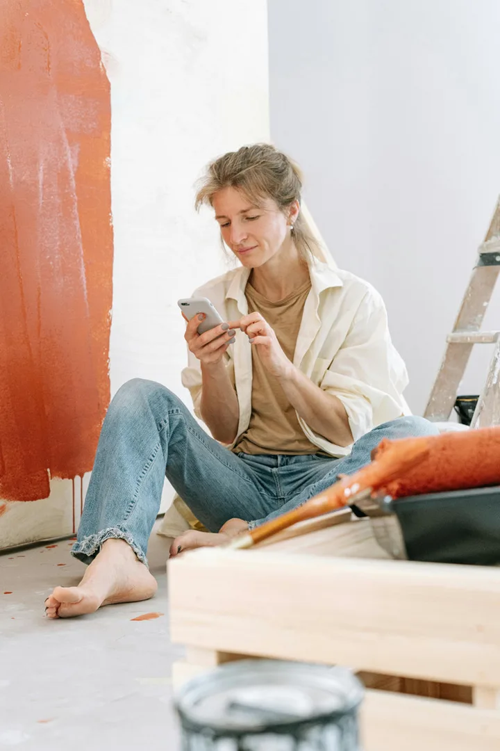 Woman sitting on floor using phone beside a red-painted wall with ladder and paint supplies nearby.