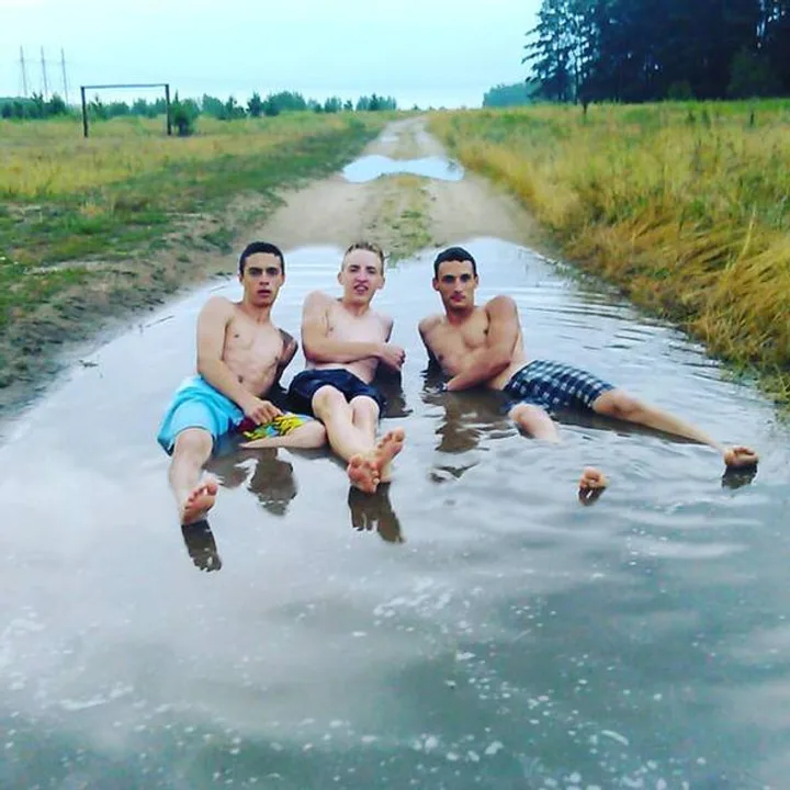 Three shirtless young men sitting and laughing in a flooded rural dirt road puddle.