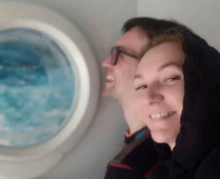 A smiling couple pressed together peering out of an airplane porthole window.