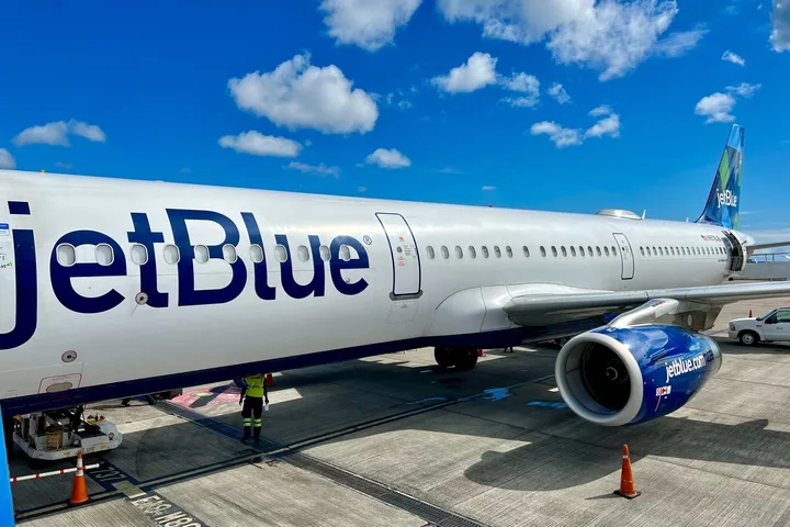 Close-up of JetBlue aircraft fuselage at gate under blue sky with ground crew visible.