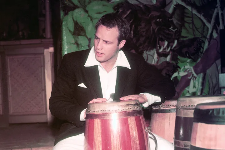 Young man in a black jacket playing bongo drums in front of a painted jungle mural.