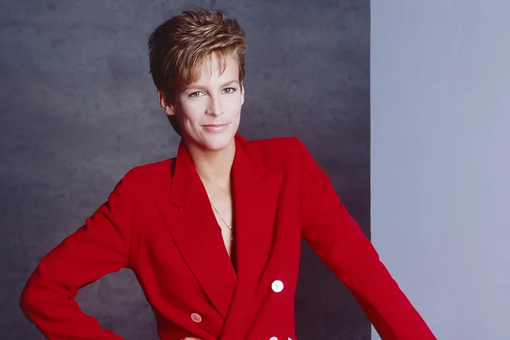 Woman with short hair wearing a bold red double-breasted blazer, posing confidently against a grey background.