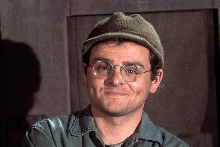 Man wearing round glasses and a knit cap smiling warmly in a dimly lit indoor setting.
