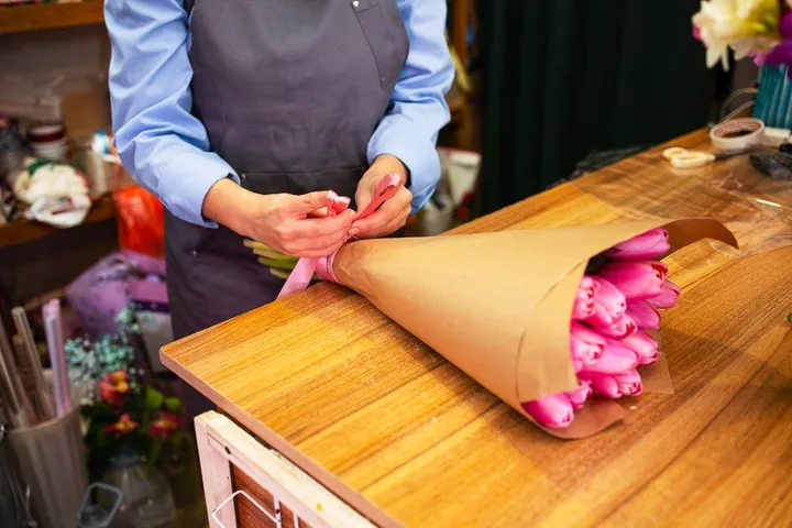 Florist in apron wrapping a bouquet of pink tulips in kraft paper at a shop counter.
