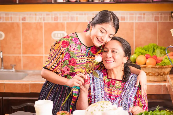 Young woman and older woman in colorful traditional embroidered dresses embracing warmly in a kitchen.