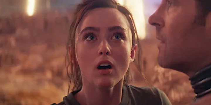Young woman with wide eyes looking shocked in a fiery, apocalyptic setting beside a man.