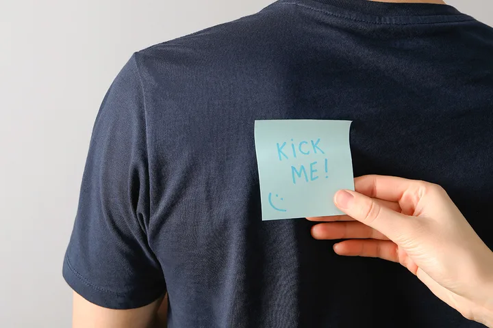 Hand secretly sticking a 'Kick Me!' sticky note on the back of someone's shirt.