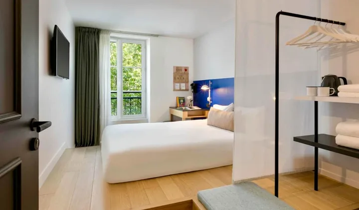 Modern, minimalist hotel room with white bed, blue headboard, and large window overlooking greenery.