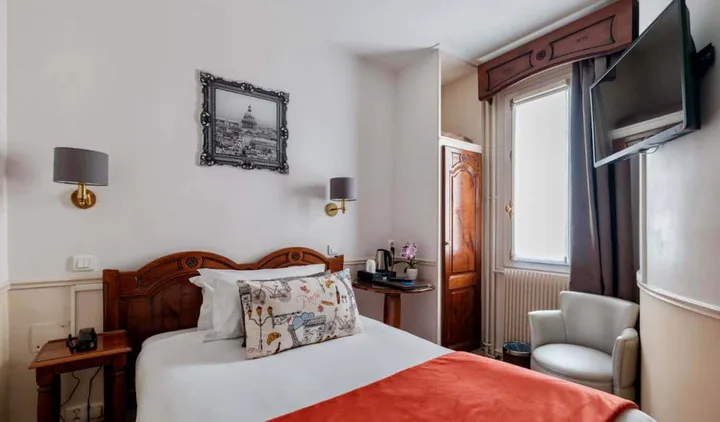 Classic hotel room with wooden headboard, red bedspread, wall sconces, and a Paris print above the bed.