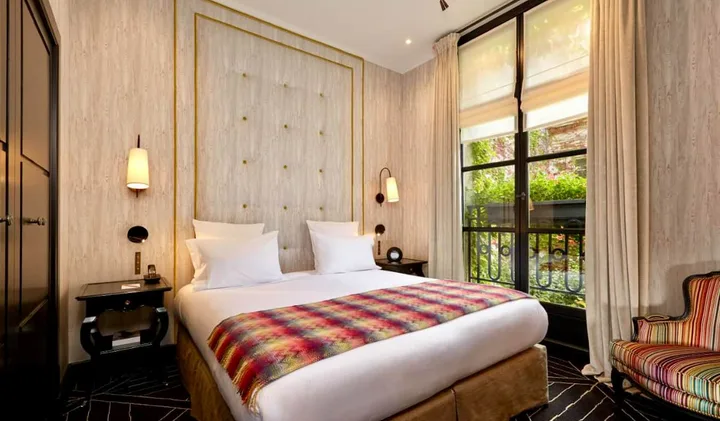 Elegant hotel room with tufted headboard, colorful throw blanket, and French doors opening to a garden balcony.