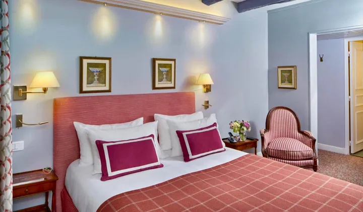 Bright hotel room with pink upholstered headboard, matching armchair, and framed art on blue-grey walls.