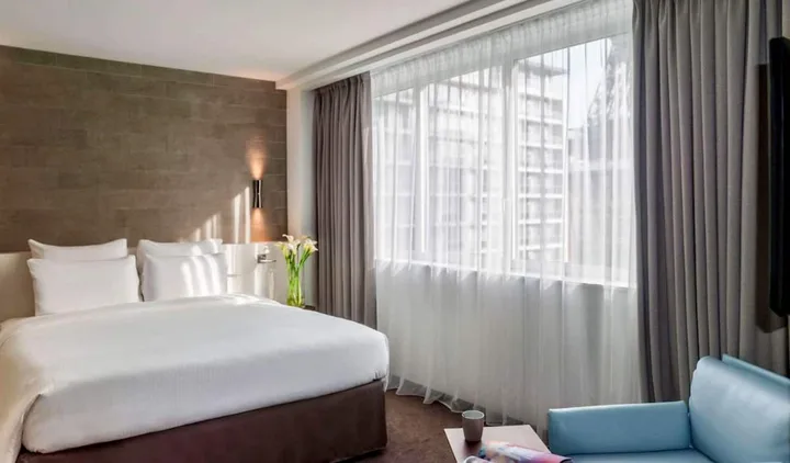 Contemporary hotel room with large sheer windows, white bed, grey curtains, and a blue armchair.