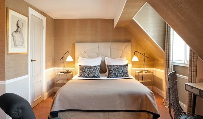 Cozy attic-style hotel room with sloped ceiling, warm grasscloth walls, and elegant neutral bedding.