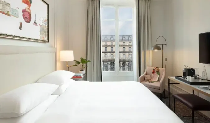 Bright hotel room with white bedding, Parisian-view balcony window, art print, and modern furnishings.