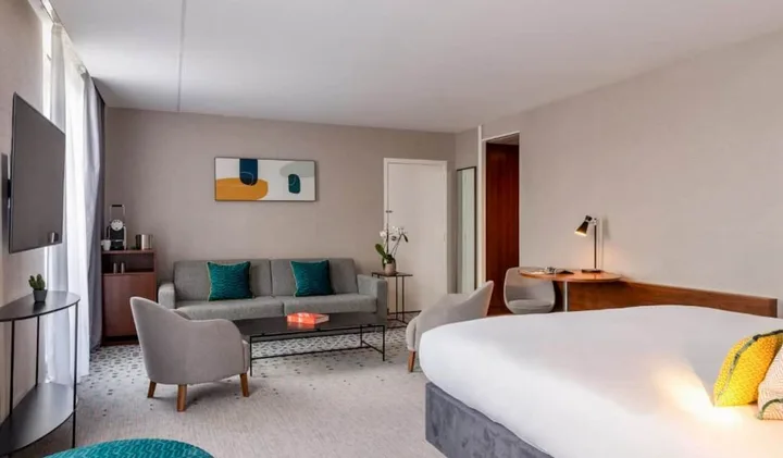 Spacious hotel junior suite with grey sofa seating area, teal accents, and modern artwork above.
