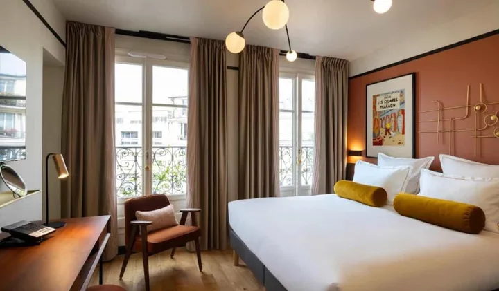Stylish hotel room with terracotta accent wall, vintage poster, Haussmann windows, and warm lighting.