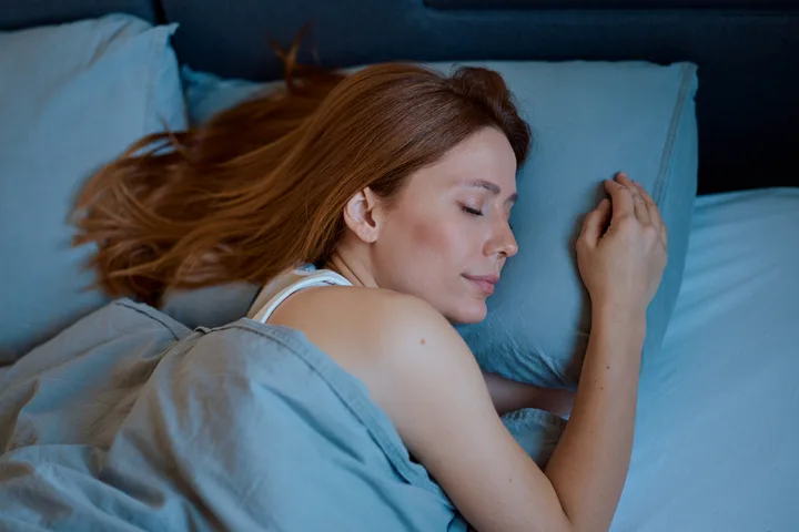 Red-haired woman peacefully sleeping in bed with blue sheets in a dimly lit room.