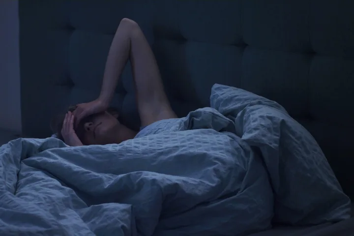 Person lying awake in a dark bedroom with hands over their face, looking distressed.