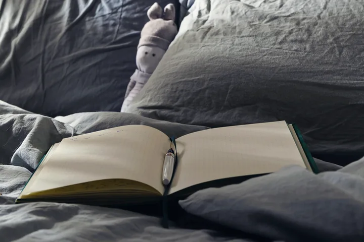 Open blank journal with a pen resting on it, sitting on rumpled grey bed sheets.
