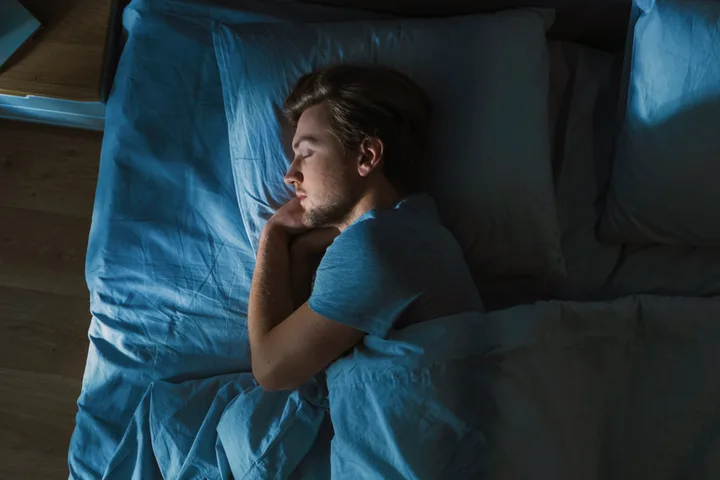 Young man sleeping peacefully in bed with blue sheets, viewed from above in dim light.