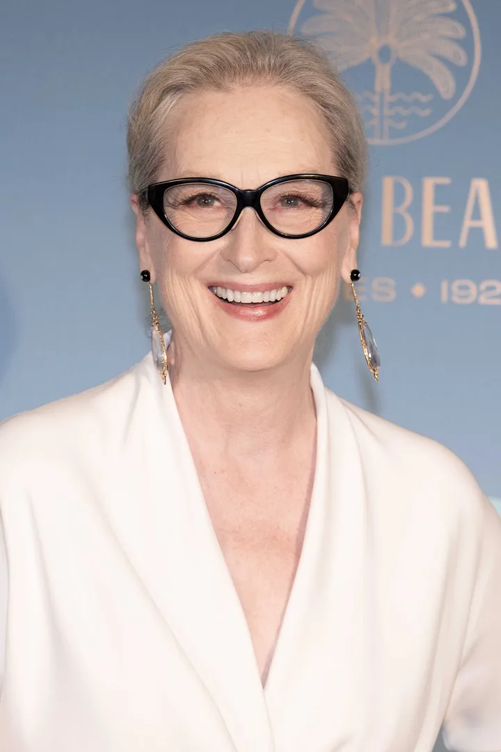 Smiling older woman with cat-eye glasses and white blazer at a formal event.