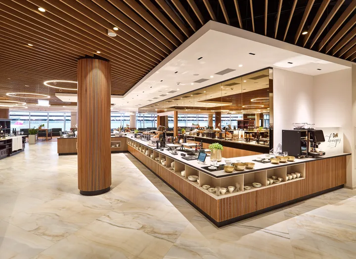 A modern, upscale airport lounge interior with wooden accents, buffet counter, and espresso machine.
