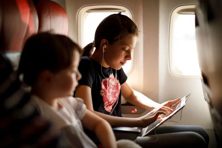 Two young girls seated in economy class, one using a tablet with earphones during a flight.