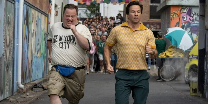 Two men running through a graffiti-lined alley with a crowd behind them in a movie scene.