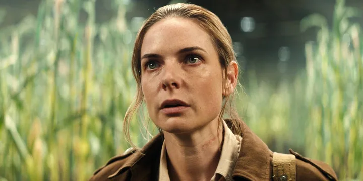 Woman with tense expression standing in a misty cornfield, wearing a brown jacket in a cinematic scene.