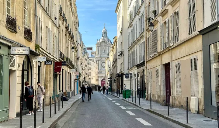 Quiet Parisian street lined with Haussmann buildings, shops, and a church dome in the background.