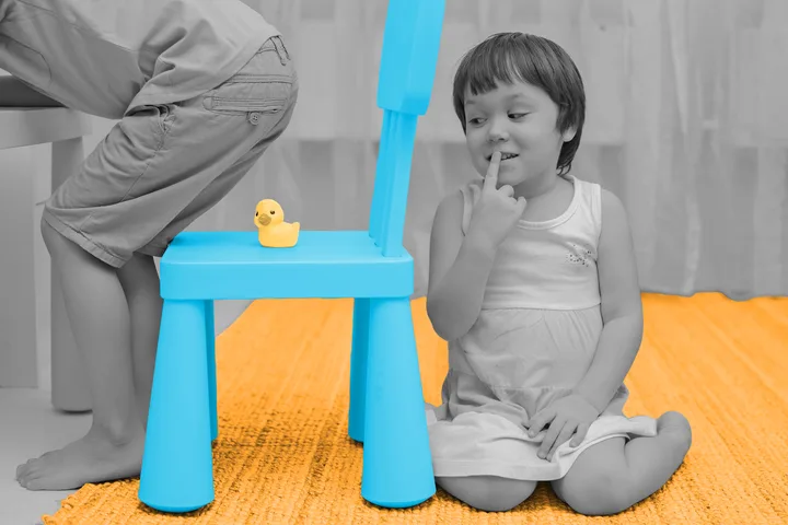 Young child sitting on floor looking at rubber duck on a blue toy chair, playful mood.