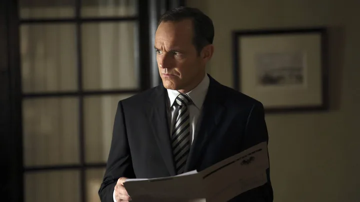 A suited man holding documents stands in a dimly lit office, looking serious and composed.
